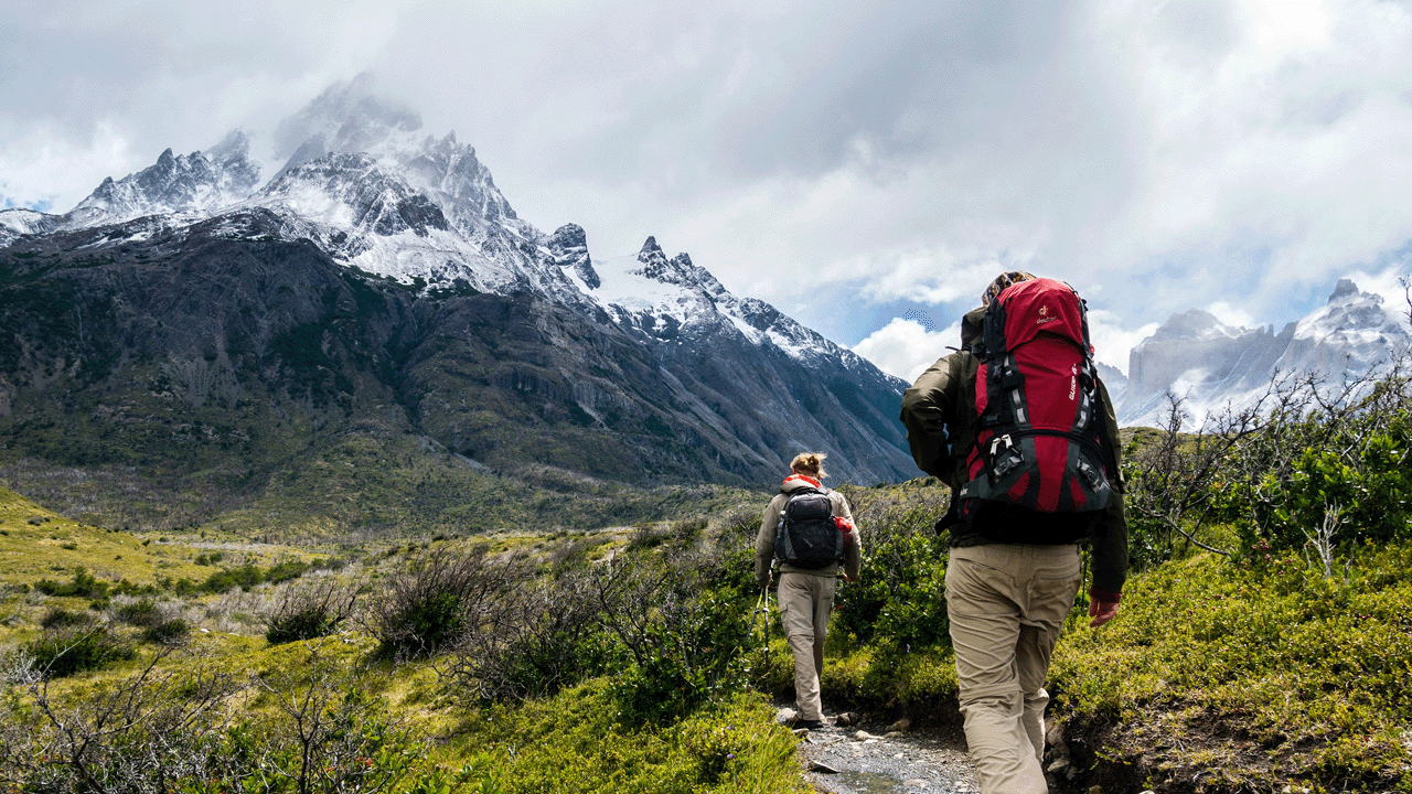 Two people carrying backpacks hiking on a trail towards a snowy mountain