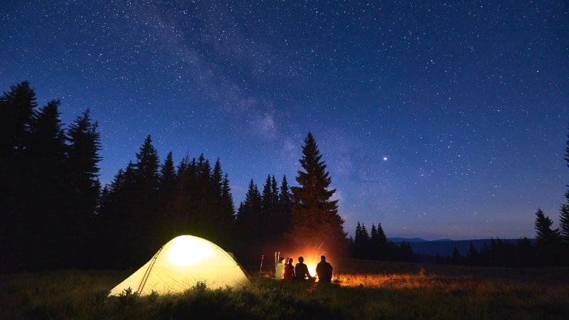 Group of people camping next to a fire with a small lit tent under the night sky